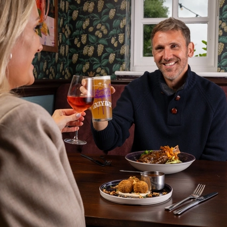 A lifestyle image focusing on a customer sat with a friend at a table celebrating with a cheers while enjoying a bowled main and plated starter dish from the January/February Specials menu with a selection of drinks within the interior restaurant seating area at a Community Pubs with Food venue.