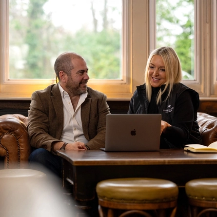 A Man and a Woman sat on a sofa. The woman is looking at the laptop on the table in front of her, The Man is looking at the Woman. 