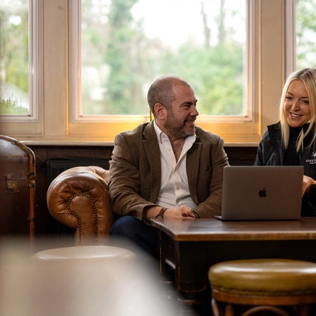 A Man and a Woman sat on a sofa. The woman is looking at the laptop on the table in front of her, The Man is looking at the Woman. 