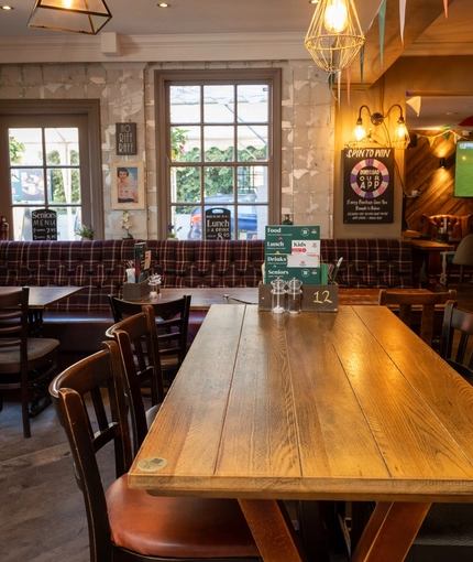 An interior restaurant seating area at The Woolpack Inn; with wooden flooring, an upholstered booth seat against the windows, and large TVs on the walls.
