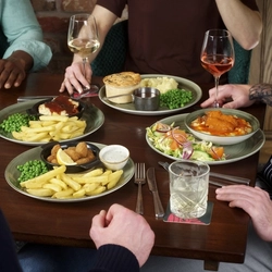 A round table filled with main dishes, including scampi and chips and a pie with mash and peas. Several drinks are placed around the table. The hands and arms of multiple people seated together are visible as they reach toward their food and raise their drinks.