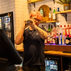 A bartender mixing a cocktail in a cocktail shaker. Plants and bottles of wine and spirits sit on shelves behind them.