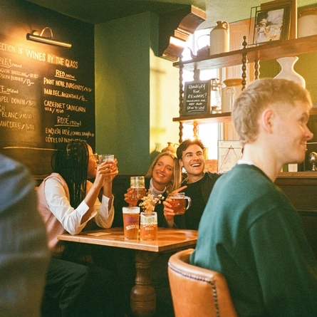 A group off friends sat in the interior restaurant and seating area in a Greene King venue in London holding drinks.