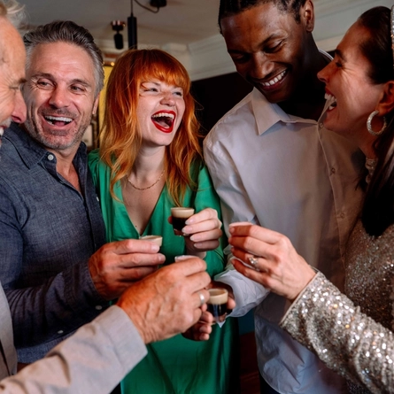 Five people gathered closely together indoors, each holding a small shot glass during a celebratory toast. One person wears a green outfit and another a silver sequined top. The group stands in a warmly lit venue, with drinks raised and decorative lighting visible in the background.