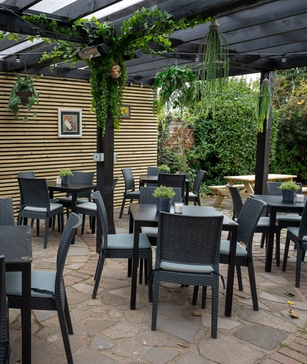 The exterior under cover beer garden seating area at The White Lion, with framed artwork on the wall, and plant baskets hanging from the wooden beams of the ceiling.