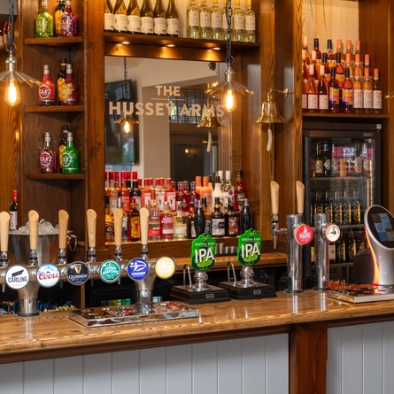 A close up view of the bar at The Hussey Arms, with silver drinks trays on the counter, and bottles of wine and alcoholic spirits on wooden shelves behind the bar.