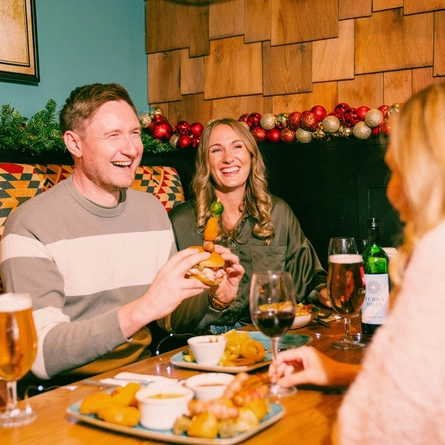 Three people sit in a festive pub booth decorated with Christmas garlands and red baubles. A wooden table is spread with sharing dishes including pigs in blankets, roast potatoes, dipping sauces and burgers, alongside glasses of lager, stout and red wine. One person takes a large bite from a stacked burger while the others enjoy food and drinks in a cosy Christmas pub setting.