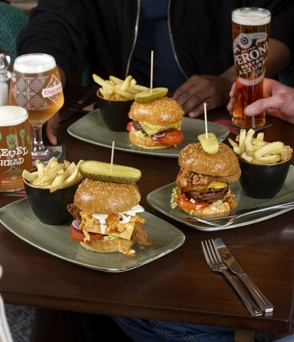 A table with three plates of burgers topped with pickles and served with fries in small cups. Several drinks—including pints of beer—are placed around the table, and the hands and arms of multiple people seated together are visible near the food.