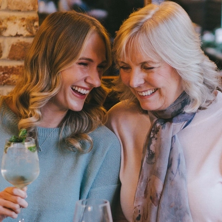 Four people sitting at a restaurant table with plates of food in front of them. One person is holding a cocktail, and a glass of wine also sits on the table.