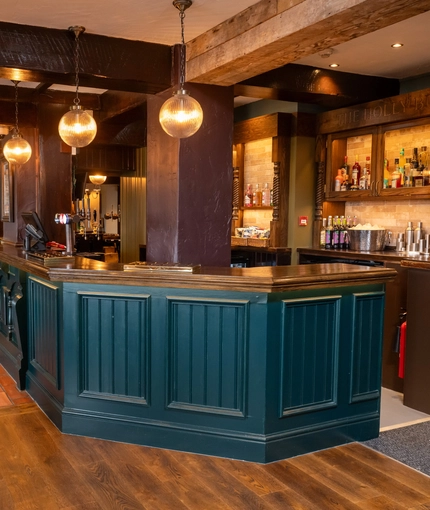 The wood panelled bar, and interior restaurant seating area of The Hollybush in Lichfield, with a fireplace at the end of the room, and four spherical glass lights hanging above the counter.