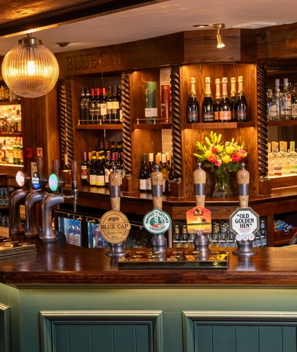 A close up view of the wood panelled bar at The Blue Cap in Sandiway, with bottles of wine and alcoholic spirits on wooden shelves behind the bar, and spherical glass lights hanging above the counter.