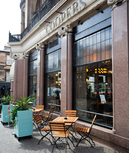 The exterior facade, signage and beer garden seating area with wooden tables and chairs at Coopers.