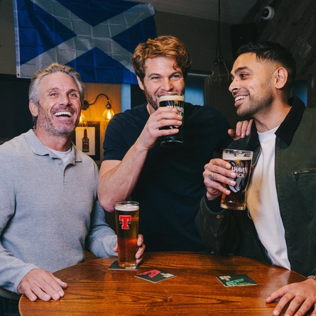 A group of friends stood round a table in the seating area in a Greene King pub with drinks watching sports.