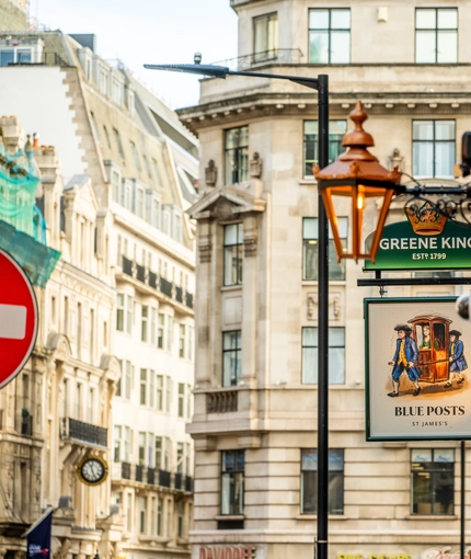 A close up view of the pub sign hanging on the exterior wall of the Blue Posts. The sign features the Greene King logo and an illustration of two people in blue coats carrying a woman in a sedan chair, above the words "Blue Posts, St James's". 