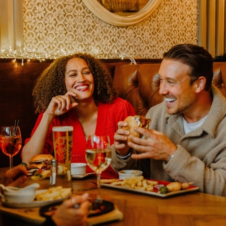 A group of diners sit in a leather booth enjoying a meal in a festively decorated pub. One diner holds a burger above a plate of chips, while drinks including wine, beer and a mixed drink are arranged on the wooden table. A small Christmas tree, warm fairy lights and a large red bow decorate the booth, creating a cosy seasonal atmosphere.