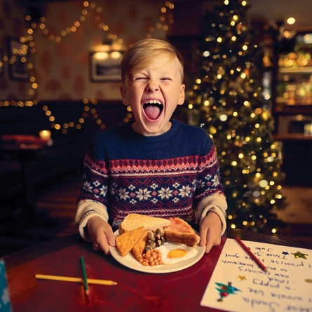A child sitting at a table inside a pub, holding a plate loaded with bread, hash browns, sausages, bacon, baked beans, mushrooms, and a fried egg. A Christmas tree and fairy lights are visible in the background.