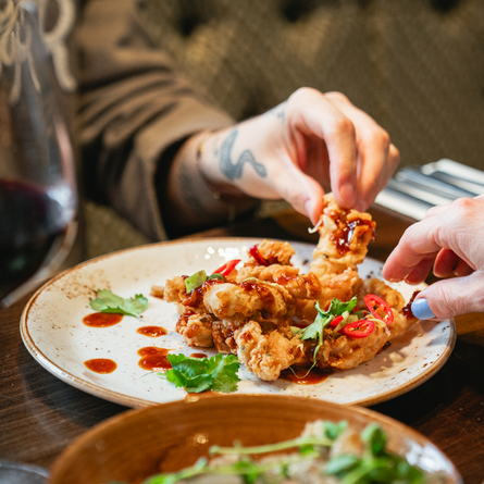 A plate of food sitting on a table next to a glass of red wine and another bowl of food. Two hands are picking up pieces of food from the plate.