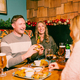 Three people sit in a festive pub booth decorated with Christmas garlands and red baubles. A wooden table is spread with sharing dishes including pigs in blankets, roast potatoes, dipping sauces and burgers, alongside glasses of lager, stout and red wine. One person takes a large bite from a stacked burger while the others enjoy food and drinks in a cosy Christmas pub setting.