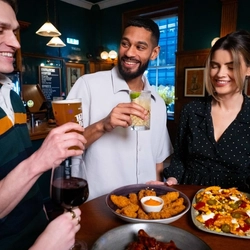 An image of a group of friends stood at a table within the restaurant area with a sharer dishes and drinks available at Urban Social venues.
