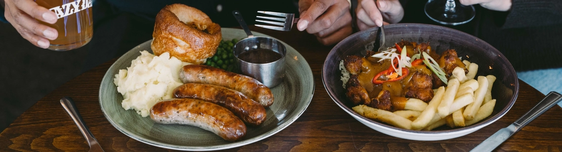 Two people seated together at a pub table with plates of sausages, mash and Yorkshire pudding, and curry with rice and chips, alongside a pint of beer and a glass of rosé.