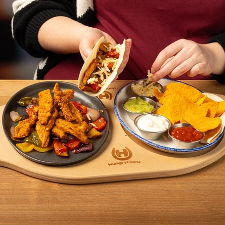 A board holding a skillet of chicken and a plate of dips, nachos and a wrap on a wooden table.  A person seated at the table is holding a fajita.
