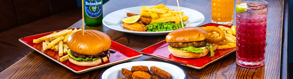 A bowl of chicken nuggets with dipping sauce, plates of burgers, chips and onion rings, glasses of drinks, and a bottle of Heineken low alcohol beer, sitting on a table.