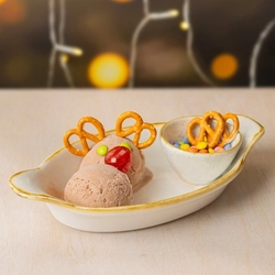 A close up view of a dish containing two scoops of ice cream and a small pot filled with sweets and pretzels. One of the scoops of ice cream is topped with two pretzels, two sweets, and a cherry to look like a reindeer; and fairy lights are visible in the background.