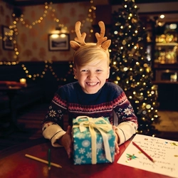 A child sitting at a table inside a pub, holding a wrapped Christmas present. A Christmas tree and fairy lights are visible in the background.