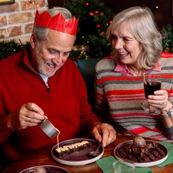 Two people seated in a festively decorated pub booth enjoy desserts and drinks at a table. One person in a red paper crown pours cream over a slice of  dessert, while the other holds a glass of mulled wine. Christmas garlands, fairy lights and a green cracker add to the seasonal setting.