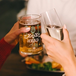 A close up view of two people sitting at a restaurant table and clinking together a glass of Level Head IPA and a glass of champagne. A burger served on a plate with a small bowl of fries, sits on the table in front of them.
