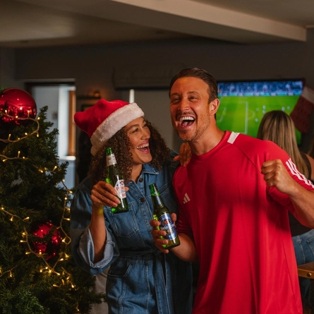A Christmas-decorated pub with a large tree adorned with red baubles, ribbon and warm fairy lights. Two people stand beside the tree holding bottled beers, one wearing a red-and-white Santa hat and the other a red sports shirt. A television in the background displays a football match while several other patrons watch with raised arms. Wooden tables and chairs are visible throughout the venue, with a plate of onion rings on a nearby table.