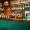 A close up view of the green, wood panelled bar inside the Phoenix; with glasses hanging in racks above the counter, and bottles of alcoholic spirits on wooden shelves behind the bar.