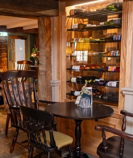 An interior restaurant seating area at the Ye Olde Leathern Bottel in Barkham, with stone flooring, wooden beams, a brick wall with a fireplace, and bottles of wine in a glass display case in another wall.