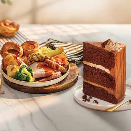 A roast dinner featuring roast meats, Yorkshire pudding, vegetables and gravy served on a plate, and a large slice of chocolate cake served on a plate with a fork. Both plates sit on a table along with a napkin and more Yorkshire puddings in the background.