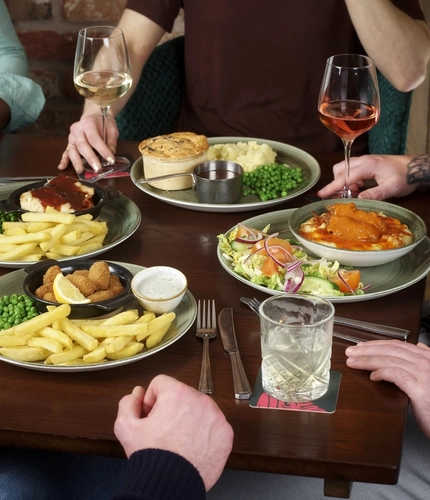 A round table filled with main dishes, including scampi and chips and a pie with mash and peas. Several drinks are placed around the table. The hands and arms of multiple people seated together are visible as they reach toward their food and raise their drinks.