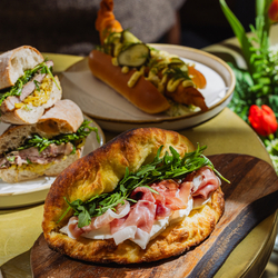 An advertising image showing 2 plated sandwich dishes and a sandwich dish presented on a wooden board sat on a table within the interior restaurant seating area at The Four Oaks.