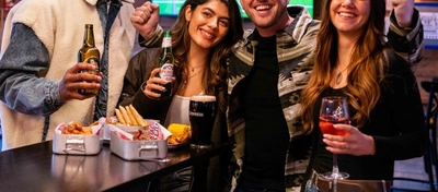 A group of people holding drinks and sitting at a table with bar snacks, posing for a photo together.