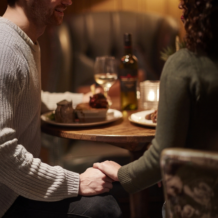Two people sat at a table holding hands lower than the table. Low lighting, you can't see the peoples faces.