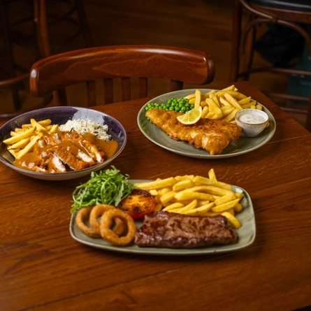 A close up view of a wooden restaurant table laden with a bowl of Katsu Chicken with fries and rice, a plate of Fish & Chips served with peas, lemon, and dipping sauce, and a plate of steak, chips, onions rings, tomato, and salad.