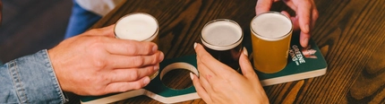 A Beer Flight Paddle is on an indoor wooden table.  There are the hands of three people seated at the table, each taking a mini pint glass of beer from it.
