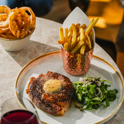 An advertising image showing a plated steak dish sat on a table with a glass of red wine at a Crafted venue.