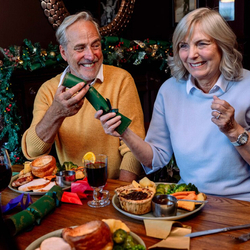 An image focusing on 2 people sat with others at a table within the interior restaurant seating area pulling a cracker while enjoying drinks and food dishes from the Festive Fayre menu at a Community Social venue.