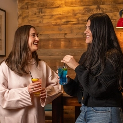 Two people leaning against a wooden table inside a restaurant, each of them holding a cocktail.