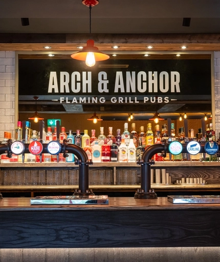 A close up view of the bar inside the Arch & Anchor, with bottles of different alcohols on a shelf behind the bar and two TVs on the wall.