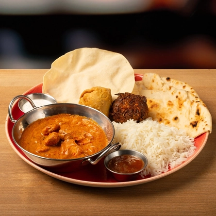 A Balti dish of curry on a plate with rice, naan bread, poppadom and a dipping sauce  on a wooden table.