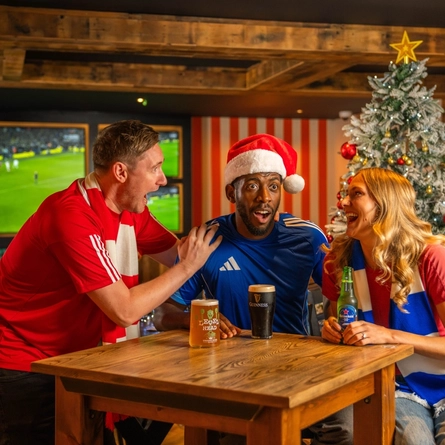 Three people gathered around a wooden table in a sports pub decorated for Christmas, watching live football on large screens. A Christmas tree stands behind them, while a pint of Guinness, a can of Level Head beer, and a bottle of Heineken 0.0 sit on the table. One person wears a Santa hat and others wear football scarves, creating a festive matchday atmosphere.