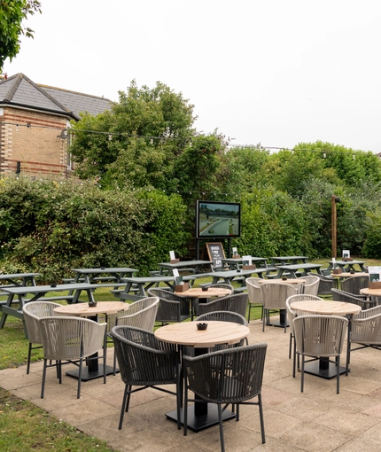 The exterior beer garden seating area at The Richmond Arms in Bournemouth, with tables and chairs on a paved patio area; wooden picnic tables on the grass; string lights overhead; and an outdoor TV.