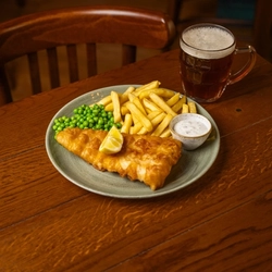 A plate of Fish & Chips with peas, lemon, and a small pot of dipping sauce. The plate sits on a wooden restaurant table along with a glass of beer.