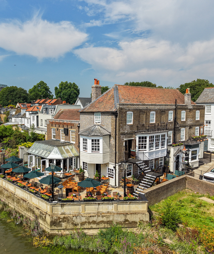An aerial view of the exterior facade, signage, and beer garden seating area at The London Apprentice in Isleworth, with a view of the waterside and surrounding buildings.