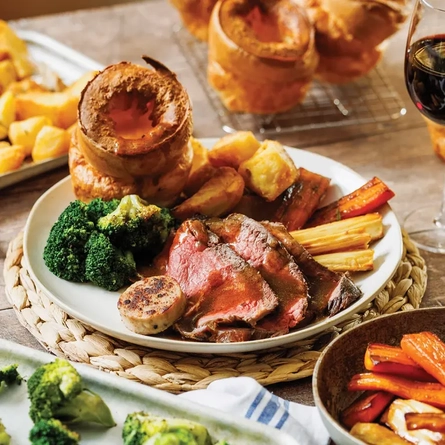 A beef roast dinner and a glass of red wine on a table surrounded by sides of vegetables.